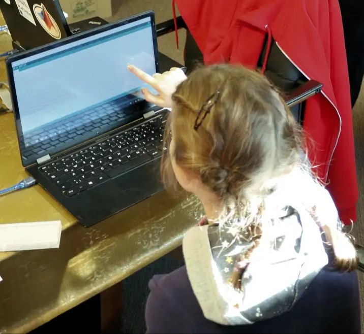 Mädchen sitzt vor einem Laptop und zeigt auf den Bildschirm.